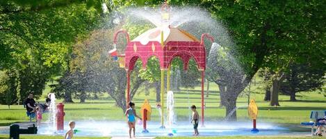 Un espace de jeux aquatiques avec une structure rouge et de nombreux jets d’eau, entouré d’herbe verte et d’arbres.
