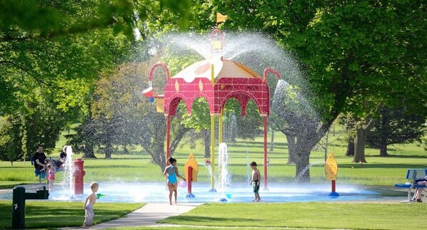 A water play area with a red structure and multiple water jets, surrounded by green grass and trees.