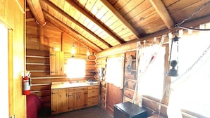 L’intérieur d’une cabane en rondins, avec un coin cuisine, des murs en bois et une fenêtre avec des rideaux.
