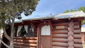 Une cabane rustique en rondins de bois, avec une porte en bois, des fenêtres avec des rideaux et une pancarte indiquant qu’il s’agit d’un lieu d’accueil intitulé « Bienvenue à la cabane ».