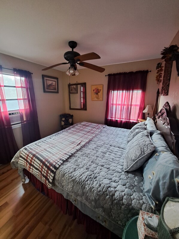 Un dormitorio con cama, ventilador de techo, ventana con cortinas, un cuadro enmarcado y espejo.