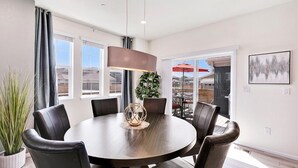 A dining area with a round table, six chairs, a pendant light, a potted plant, and a view of the outdoors.