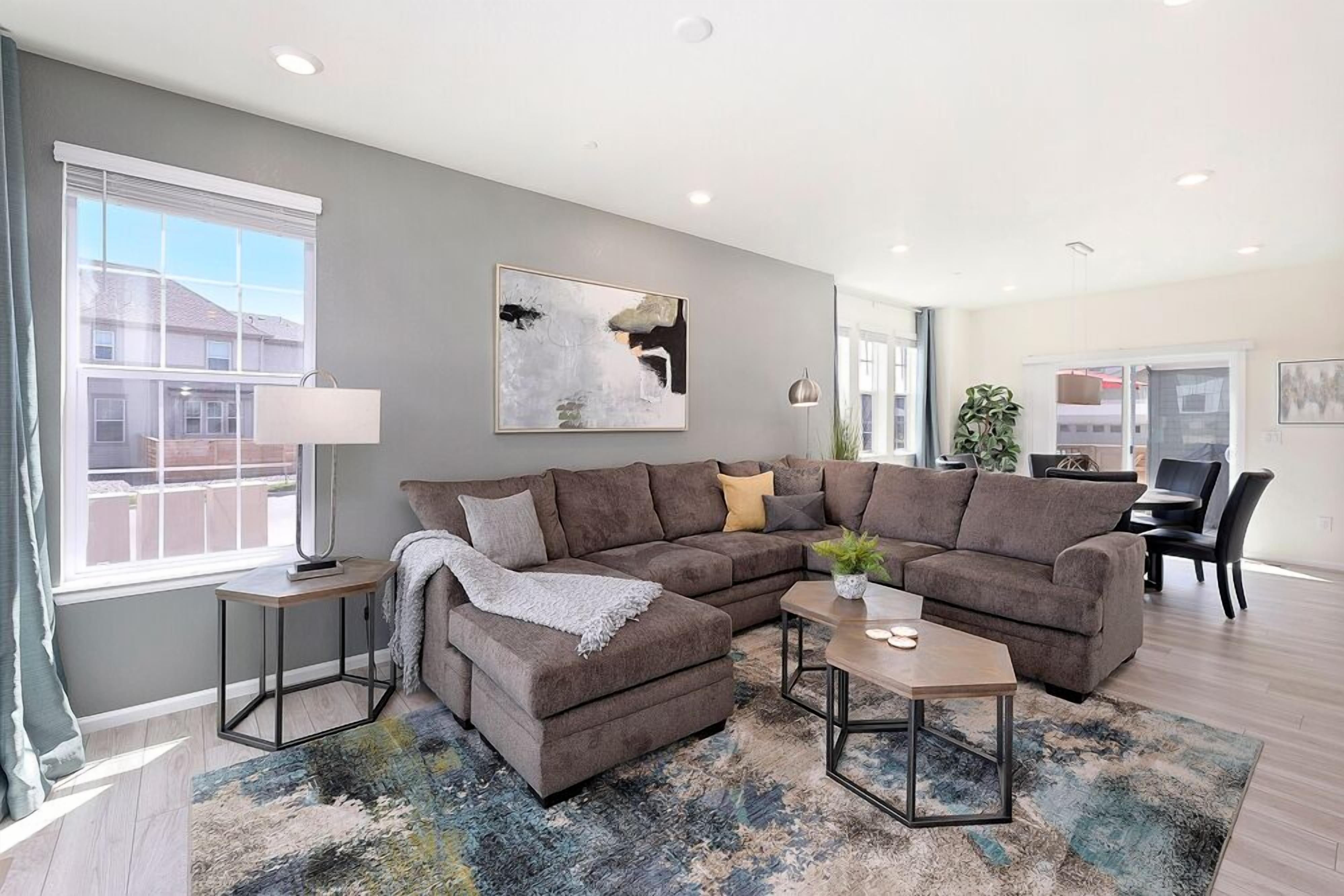 A modern living room with a sectional sofa, a coffee table, and a rug. There is a window with blinds, a lamp, and a painting on the wall.