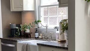 Una cocina con gabinetes blancos, encimera de granito, cafetera y ventana con vista a un edificio de ladrillo.