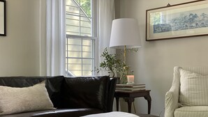 Una sala de estar con un sofá de cuero oscuro, una manta blanca, una lámpara, una mesita con libros y una vela, y un cuadro en la pared.