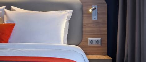 A neatly made bed with white and orange bedding, a grey headboard, and a bedside table with a light switch and power outlets.