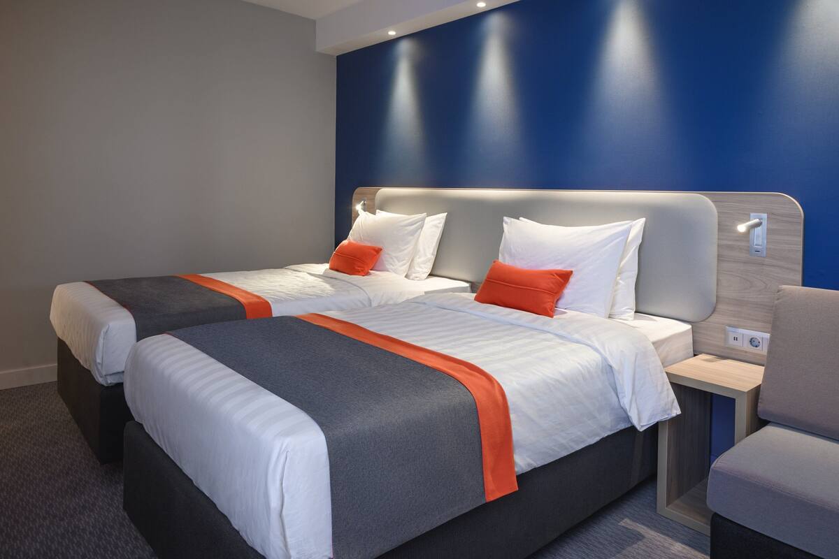 A hotel room with two beds, each with white and orange bedding, a grey headboard, and a small bedside table with a lamp.