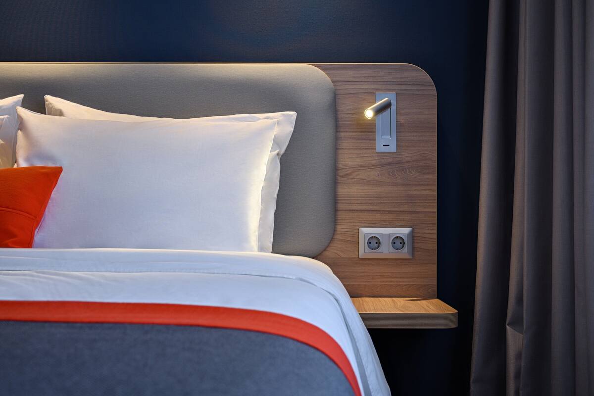 A modern hotel room with a neatly made bed, a bedside table with a lamp, and a power outlet.