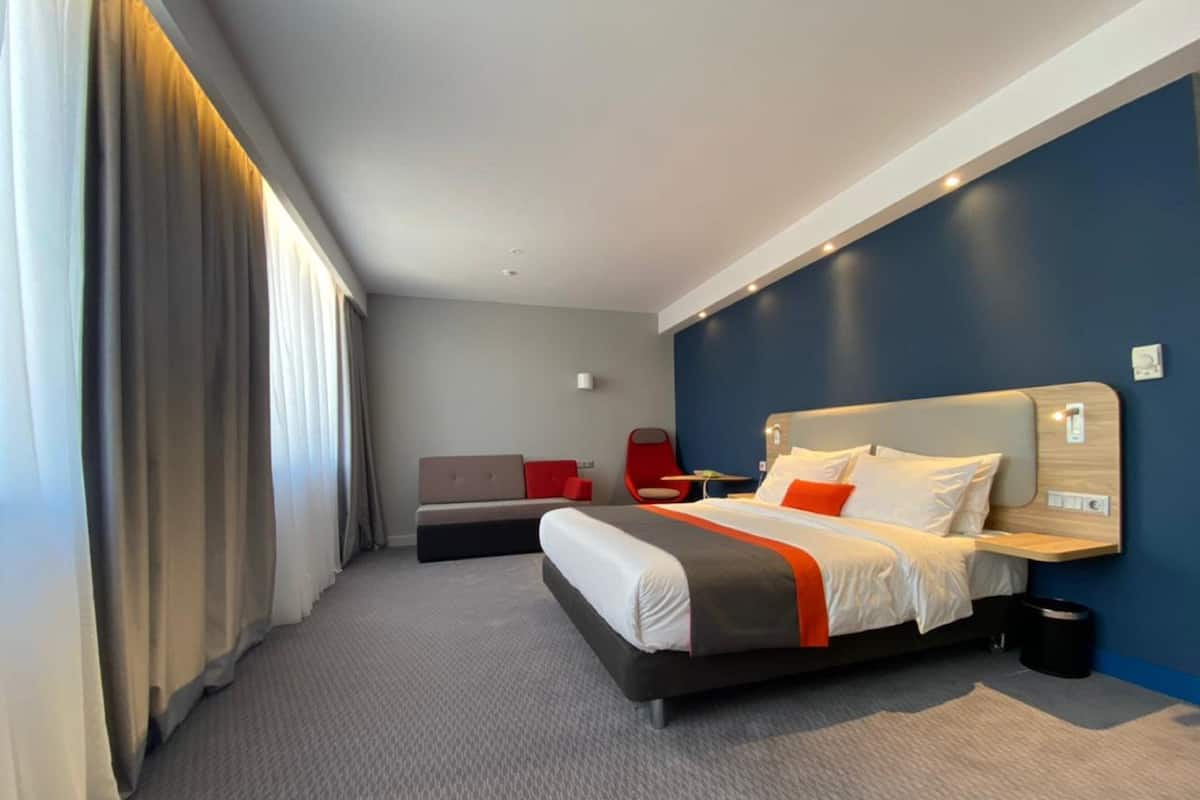 A modern hotel room with a large bed, a sofa, a red chair, and a desk.
