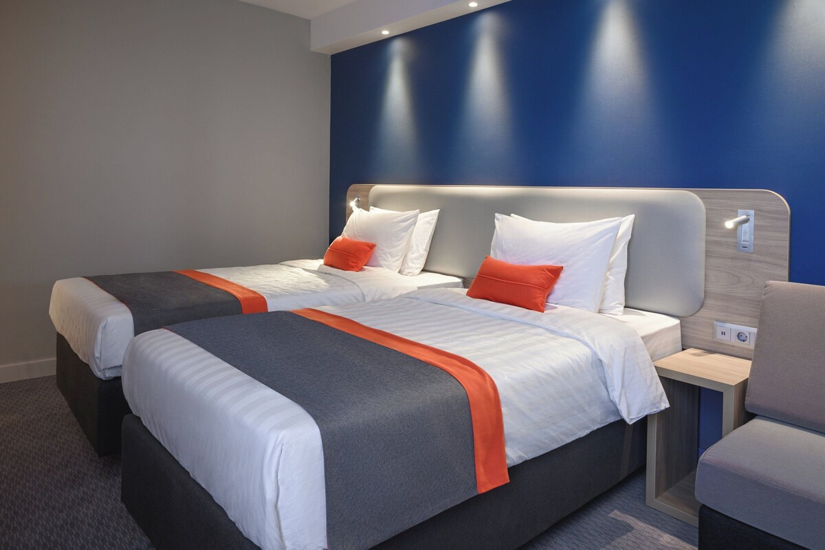 Een hotelkamer met twee bedden, elk met wit en oranje beddengoed, een grijs hoofdeinde en een klein nachtkastje met een lamp.