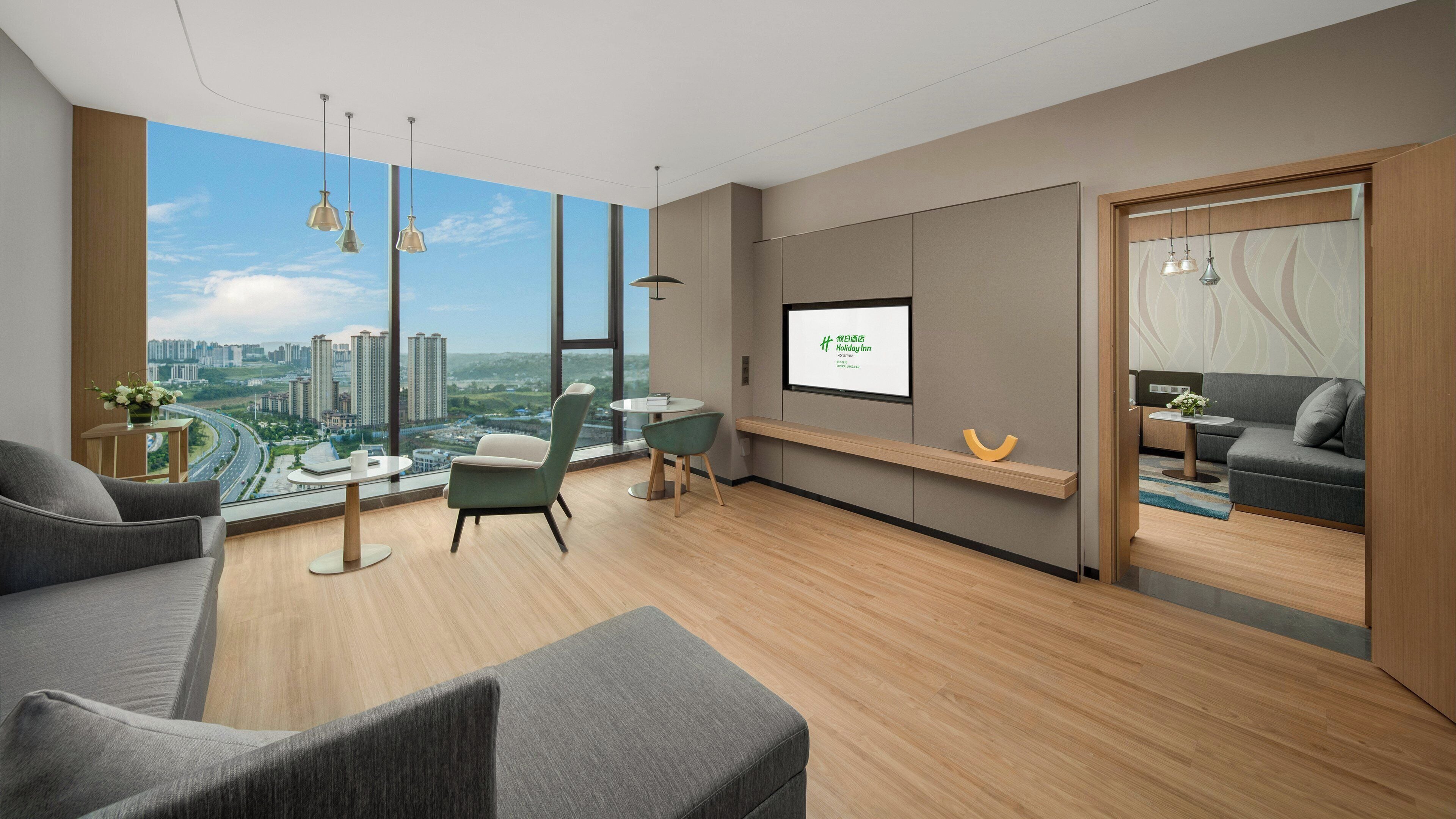 A modern hotel room with a large window, a sofa, a dining table, and a TV screen displaying the hotel name.