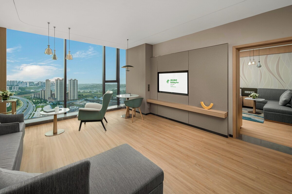 Ein modernes Hotelzimmer mit großem Fenster und Blick auf die Stadt, einem Flachbildfernseher, einer Couch, einem Esstisch und einem kleinen Balkon.