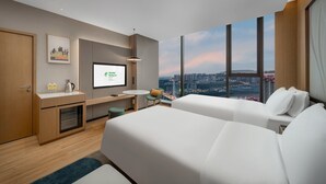 Et hotelværelse med en seng, et skrivebord med en computer, en stol, et lille bord og et stort vindue med udsigt over byen.
