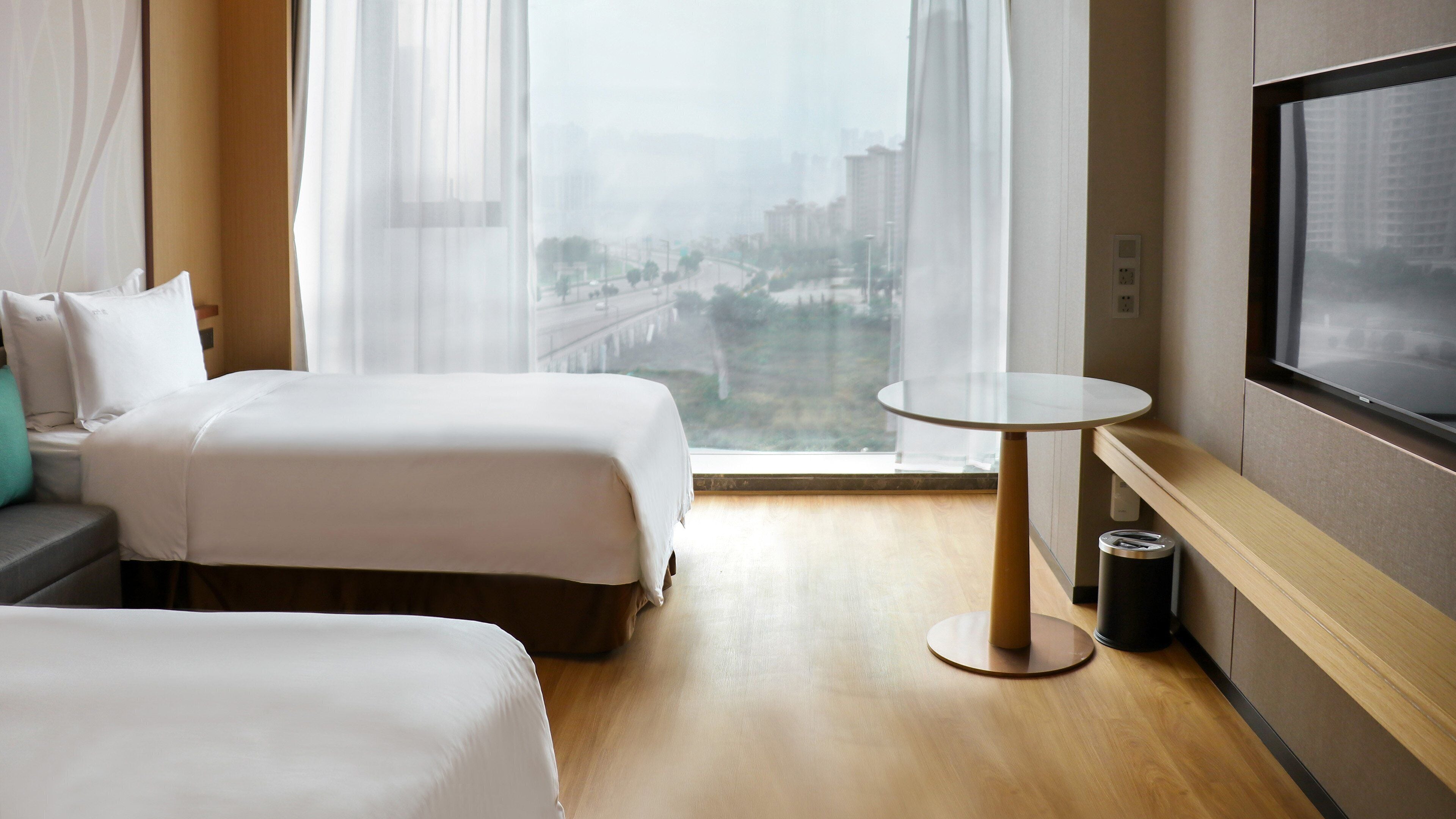 A hotel room with two beds, a small round table, a TV, and a view of a cityscape through sheer curtains.