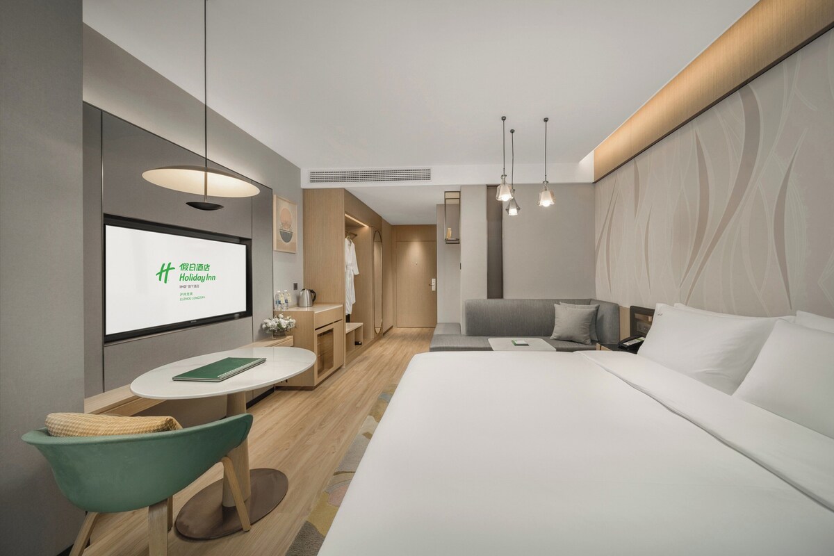 Ein modernes Hotelzimmer mit einem großen Bett, einem Schreibtisch mit Stuhl, einem kleinen Tisch und einem Fernseher, der ein Bild zeigt.