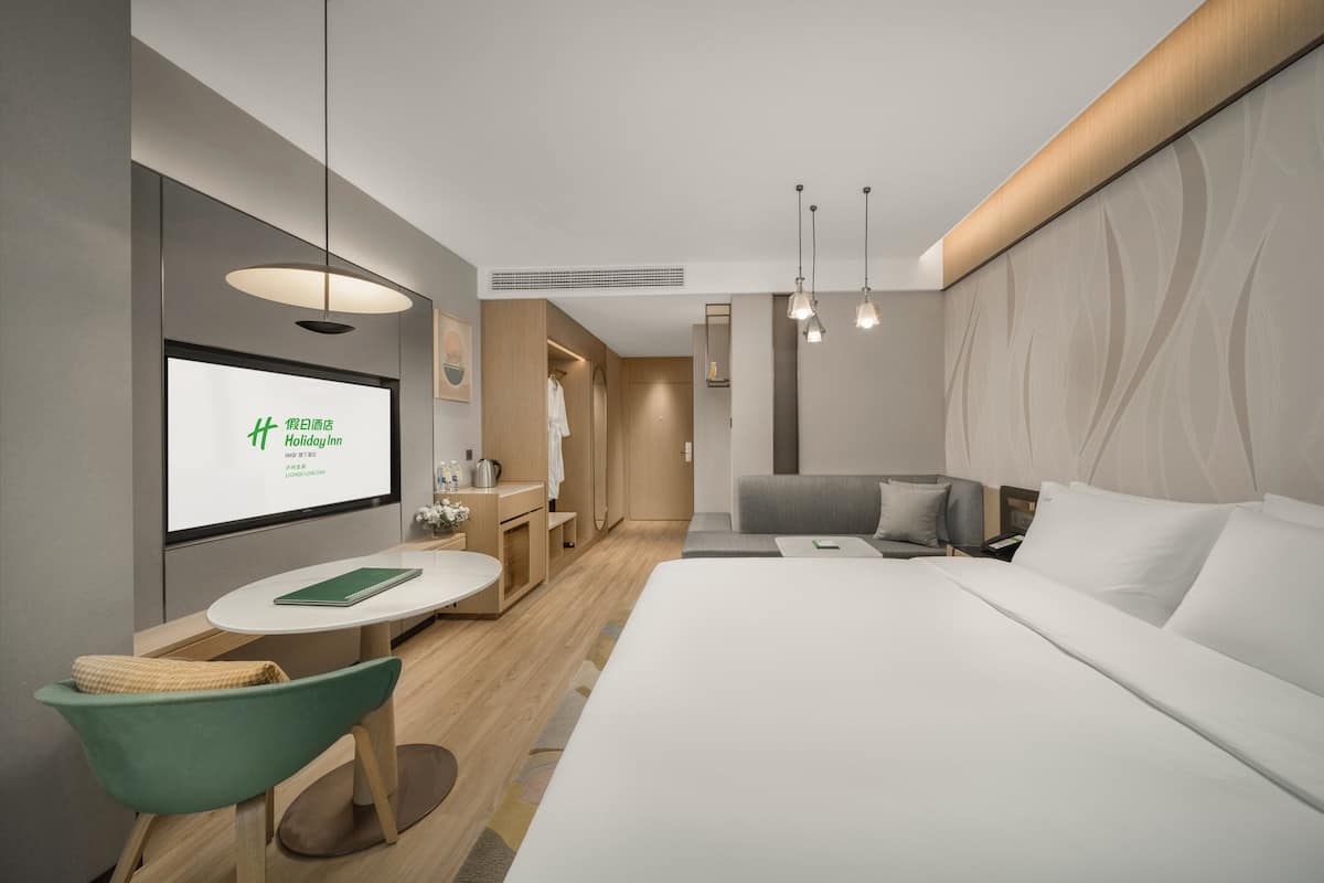 A modern hotel room with a large bed, a desk with a chair, a small table, and a TV displaying a screen.