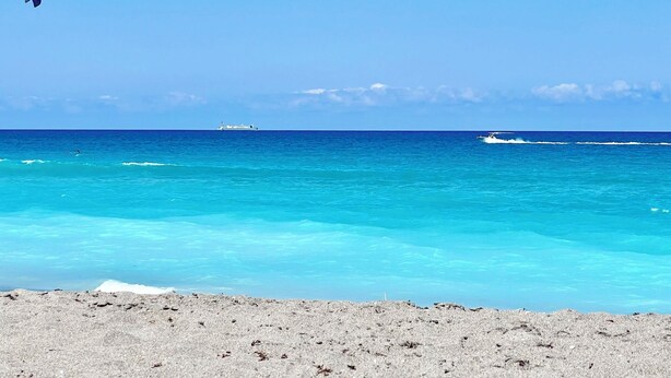 A beach with clear turquoise waters, a distant ship, and a small boat. - POSHPADZ Kai Villa-Water Views, Balcony, Walk to Jupiter Beach Sleeps 4 (Jupiter)