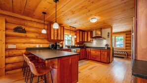 A log cabin kitchen with wooden walls, a central island with bar stools, pendant lighting, and a view of the outside. - Mountain Air, Lake Days, Fireside Nights (Swanton)
