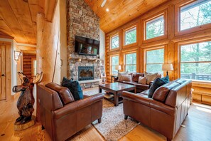 A log cabin interior with a stone fireplace, leather sofas, and large windows offering a view of the forest. - Mountain Air, Lake Days, Fireside Nights (Swanton)