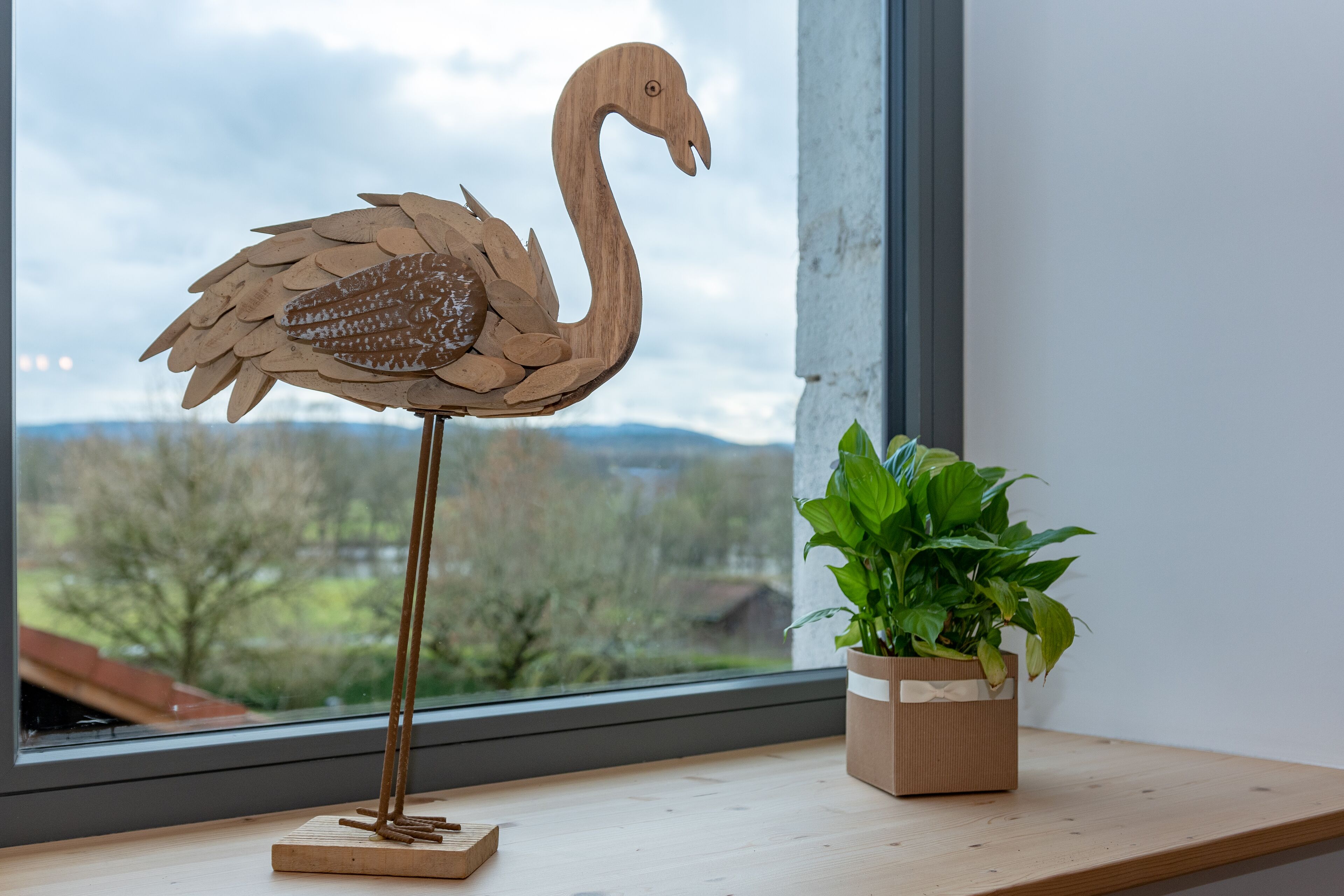 A wooden flamingo sculpture on a wooden base by a window with a view of trees and mountains.
