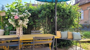 Een terras buiten met een gele tafel en stoelen, een blauwe parasol en een bloemstuk.