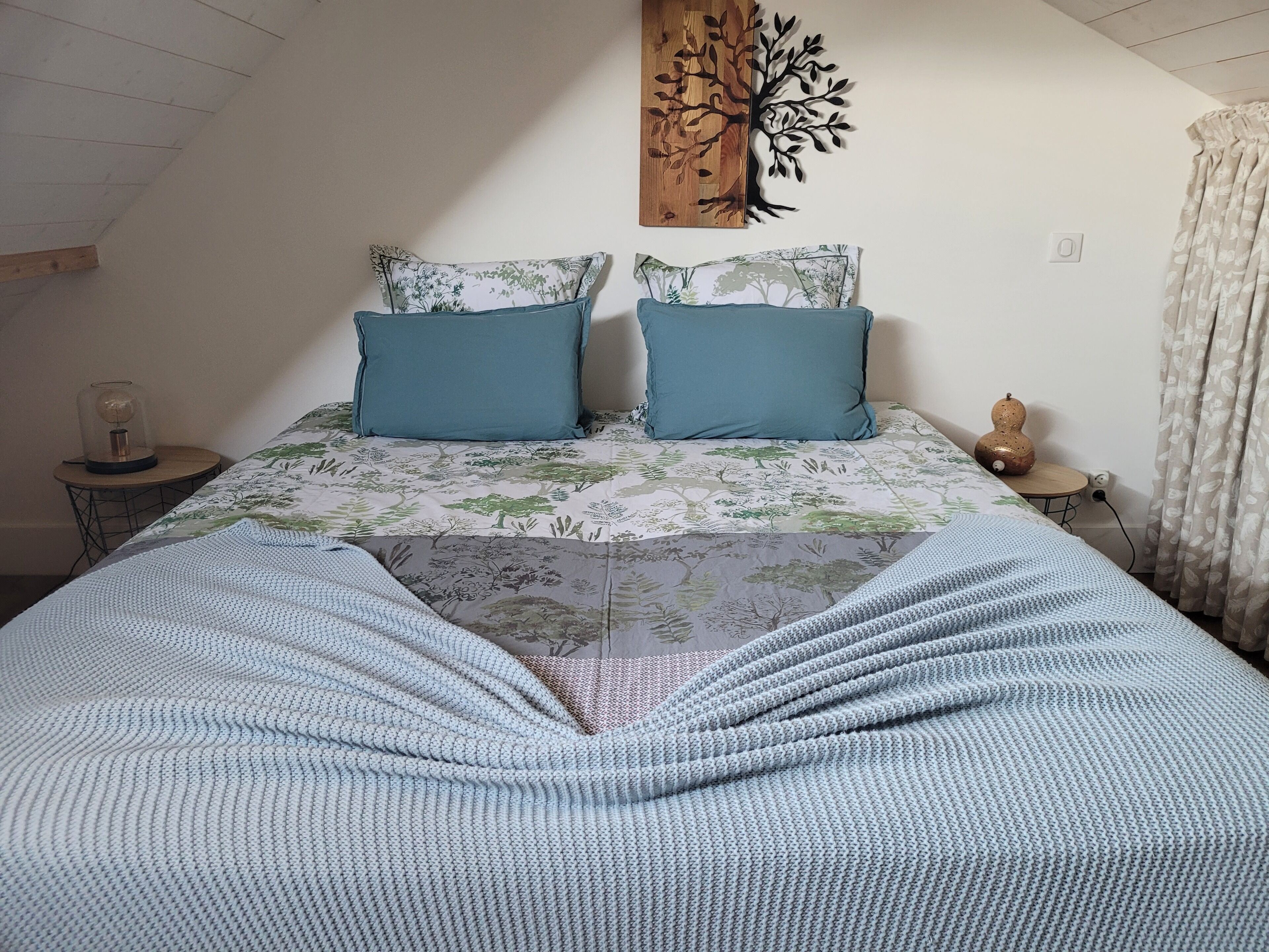 A bedroom with a bed, two pillows, a blanket, a wall art piece, a small table with a lamp, and a curtain.