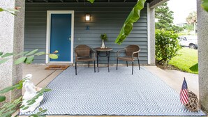 A covered porch with two wicker chairs, a small table with a plant, and a blue and white patterned rug. - Beachy Getaway–Cozy, Clean & Equipped w/ All Essentials! Relax, Enjoy & Refresh! (St. Augustine Beach)