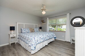 Quarto com cama, ventoinha no teto, janela com persianas, mesa de cabeceira com abajur e espelho na parede.