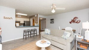 A living room with a beige sofa, a coffee table, and a kitchen in the background. - Beachy Getaway–Cozy, Clean & Equipped w/ All Essentials! Relax, Enjoy & Refresh! (St. Augustine Beach)