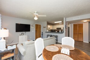 Uma sala de estar com sofá, mesa de jantar e cadeiras, uma TV fixada na parede e um ventilador de teto.
