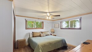 A bedroom with a bed, a ceiling fan, two windows, a bedside table, and a lamp.
