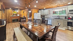 A wooden interior with a dining table, chairs, a bench, a kitchen with appliances, and a window with a view of greenery.