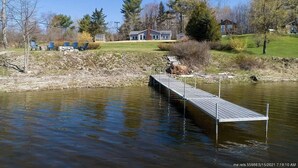 Una propiedad frente al lago con un muelle que se adentra en el agua, rodeada de árboles y una casa al fondo.