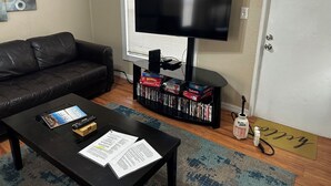 Una sala de estar con un sofá de cuero negro, una mesa de centro con papeles y un control remoto, un televisor sobre un mueble bajo, una estantería con juegos de mesa, y una puerta con felpudo.