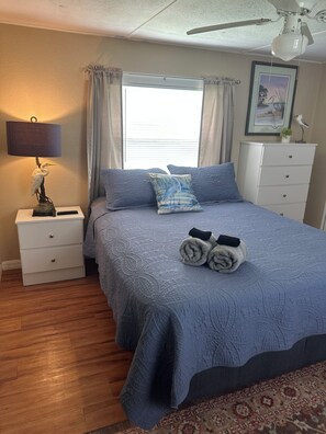 Une chambre à coucher comprenant un lit, une table de chevet, une lampe, une commode, un ventilateur de plafond et une fenêtre avec des rideaux.