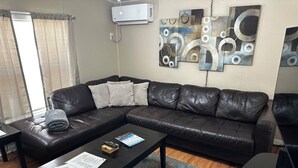 Una sala de estar con un sofá de cuero negro, una mesa de centro con papeles y un control remoto, un tapete azul estampado y arte abstracto en la pared.