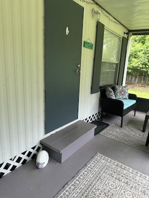 Un portico coperto con una porta verde scuro, un recinto in legno bianco, una panchina con cuscini decorati e una statuina di una pecora.
