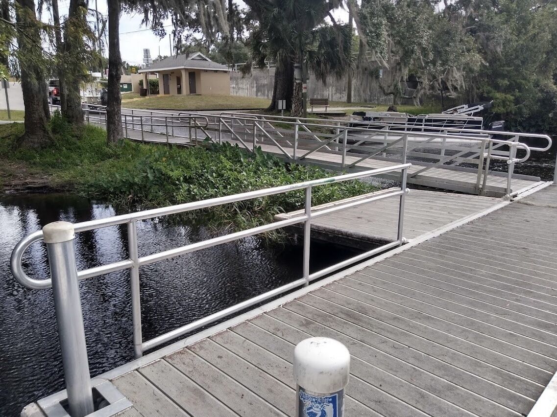 A wooden boardwalk with metal railings over a water channel, surrounded by trees and a building in the background.