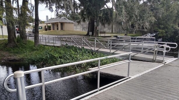 Un muelle de madera con barandillas metálicas sobre un canal, rodeado de árboles y un edificio al fondo.