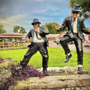 Two statues of men in business attire, one holding a microphone, positioned on a stone ledge with grass and purple foliage around. - River Retreats/Anglers Paradise (Dunnellon)