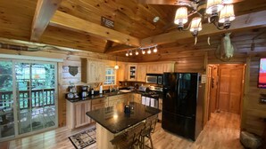 A wooden cabin interior with a kitchen, dining area, and a large window overlooking a deck.