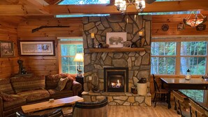 A cozy cabin interior with a stone fireplace, wooden furniture, and large windows offering a view of the forest.