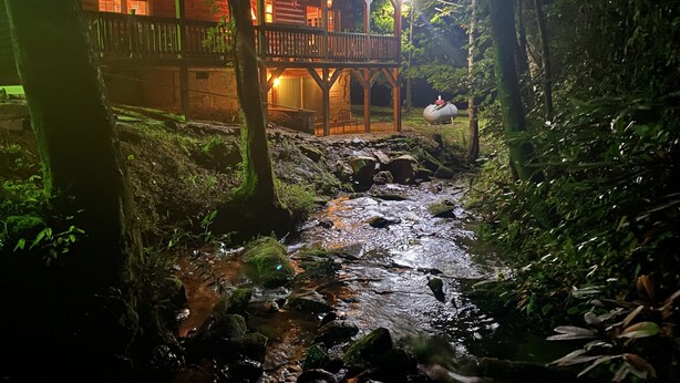 A wooden cabin with a balcony overlooking a stream, surrounded by dense forest.