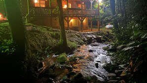 A wooden cabin with a balcony overlooking a stream, surrounded by dense forest.