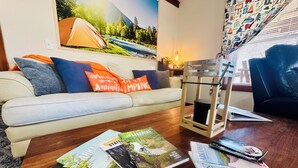 Una sala de estar con un sofá, una mesa de centro con revistas, una tienda de campaña en la pared y una ventana con persianas.