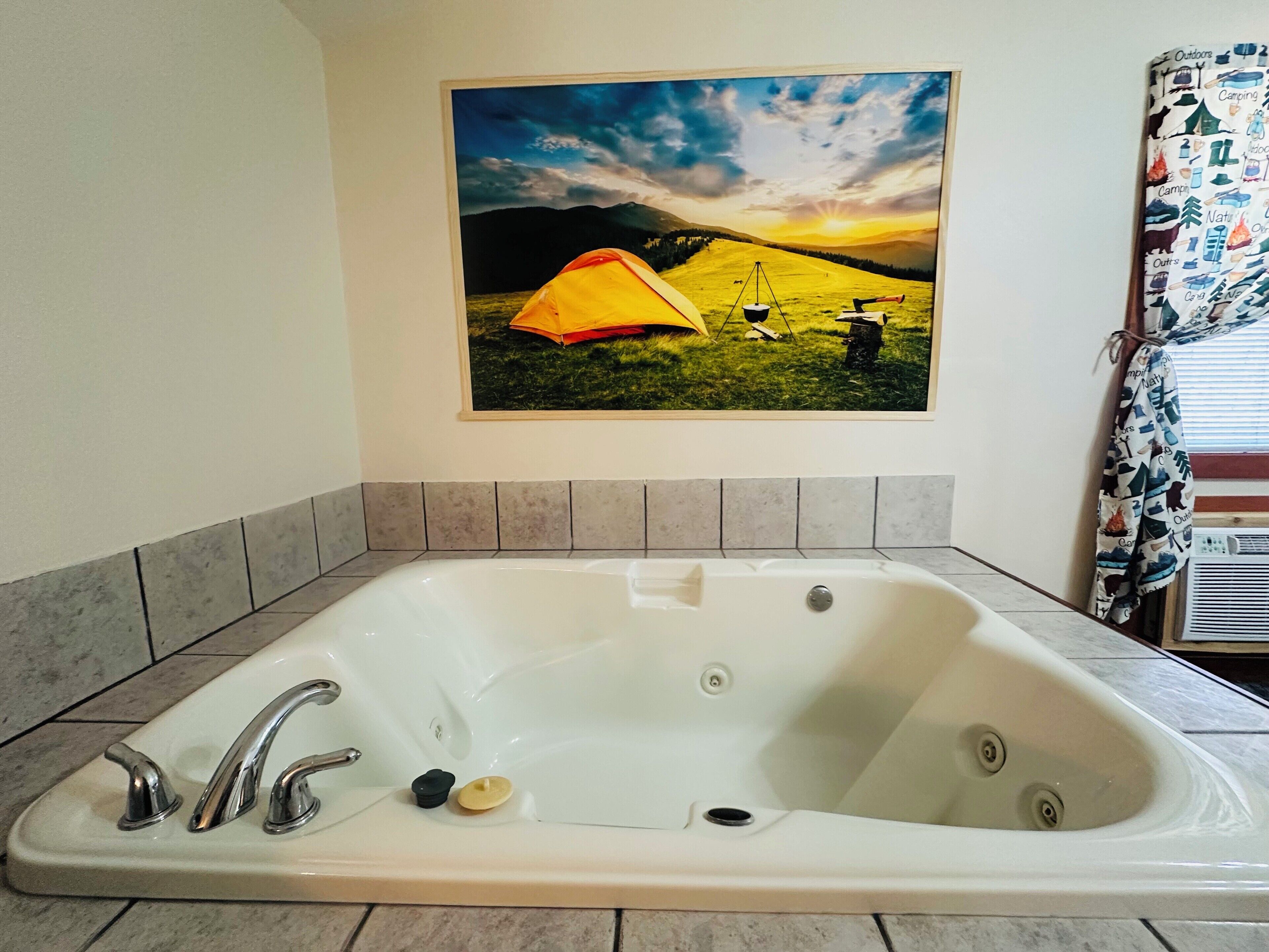 Un baño con una bañera blanca grande, un cuadro con una escena de campamento y una cortina de ducha estampada.