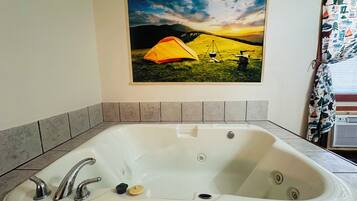 Un baño con una bañera blanca grande, un cuadro con una escena de campamento y una cortina de ducha estampada.