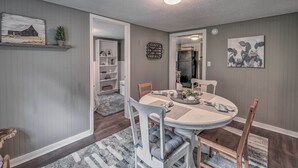 Un comedor con una mesa redonda, sillas y alfombra. Hay estantes con objetos decorativos y una puerta que da a otra habitación.