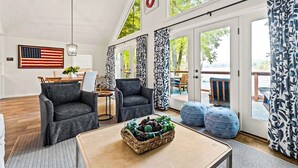 A spacious living room with two grey armchairs, a coffee table with a basket of pinecones, and a large American flag on the wall. - Bluebird Hideaway (Queensbury)