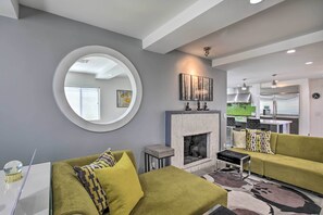 Un salon moderne avec une cheminée, des canapés verts, un miroir rond et une cuisine en arrière-plan.